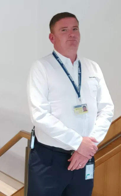 Sodexo security colleague wearing a white uniform shirt and staff lanyard standing beside an indoor staircase.