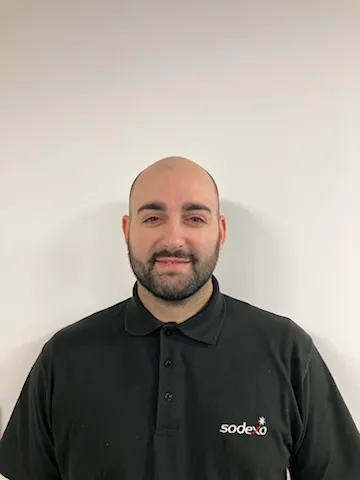 Sodexo colleague wearing a black Sodexo polo shirt standing against a plain white wall.