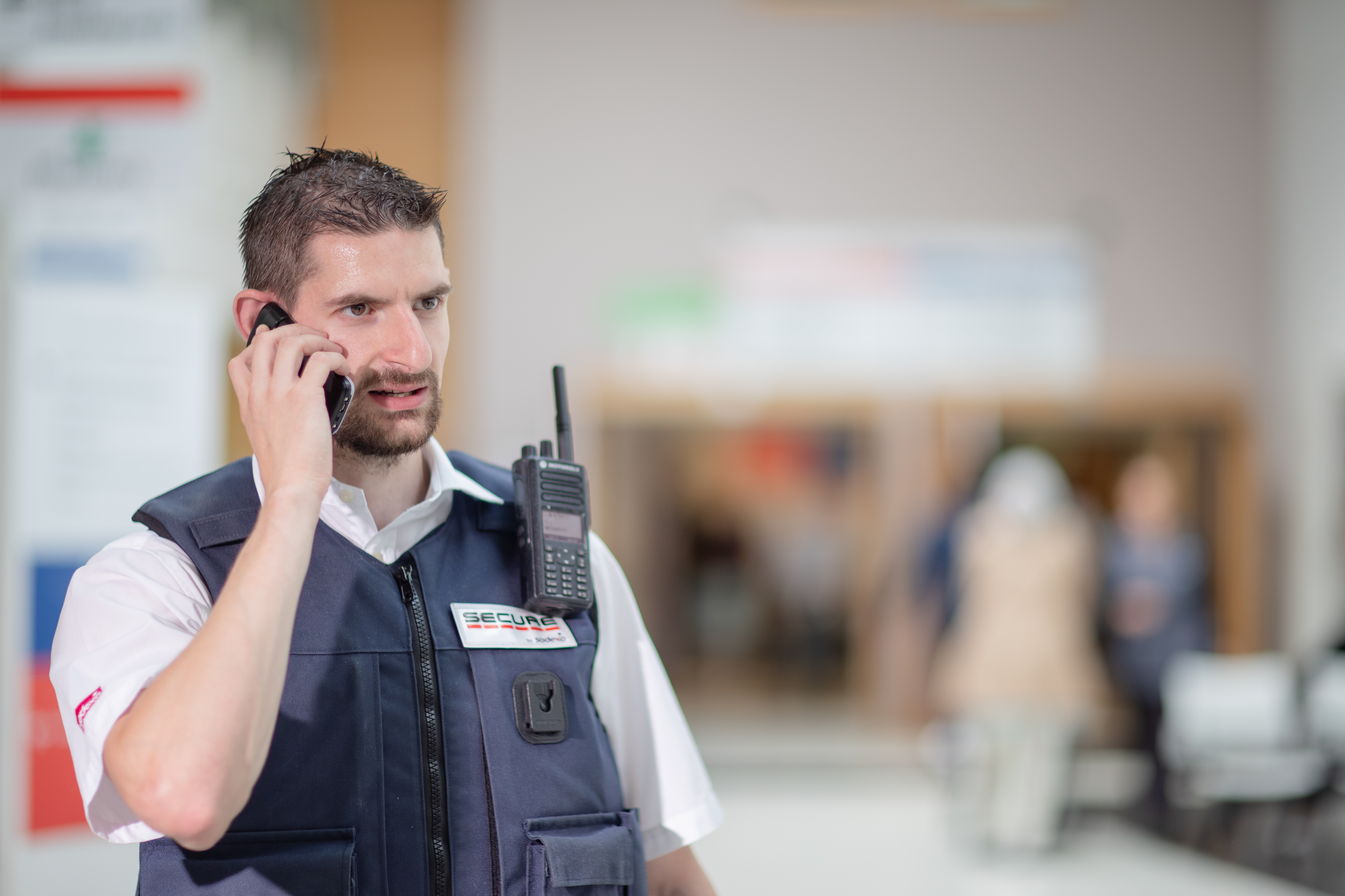 Security guard speaking on a phone while wearing a uniform and radio in a public indoor space, with blurred people and seating in the background.