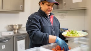 A female staff member is serving up some lettuce and tomatoes in the kitchen for a patient.