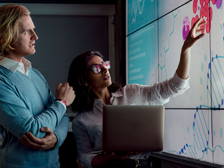 Two individuals analyzing data on a big screen displaying various graphs