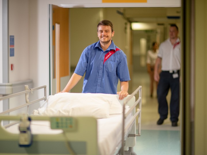 A porter pushing a trolley and smiling, there’s another porter in the background walking behind him