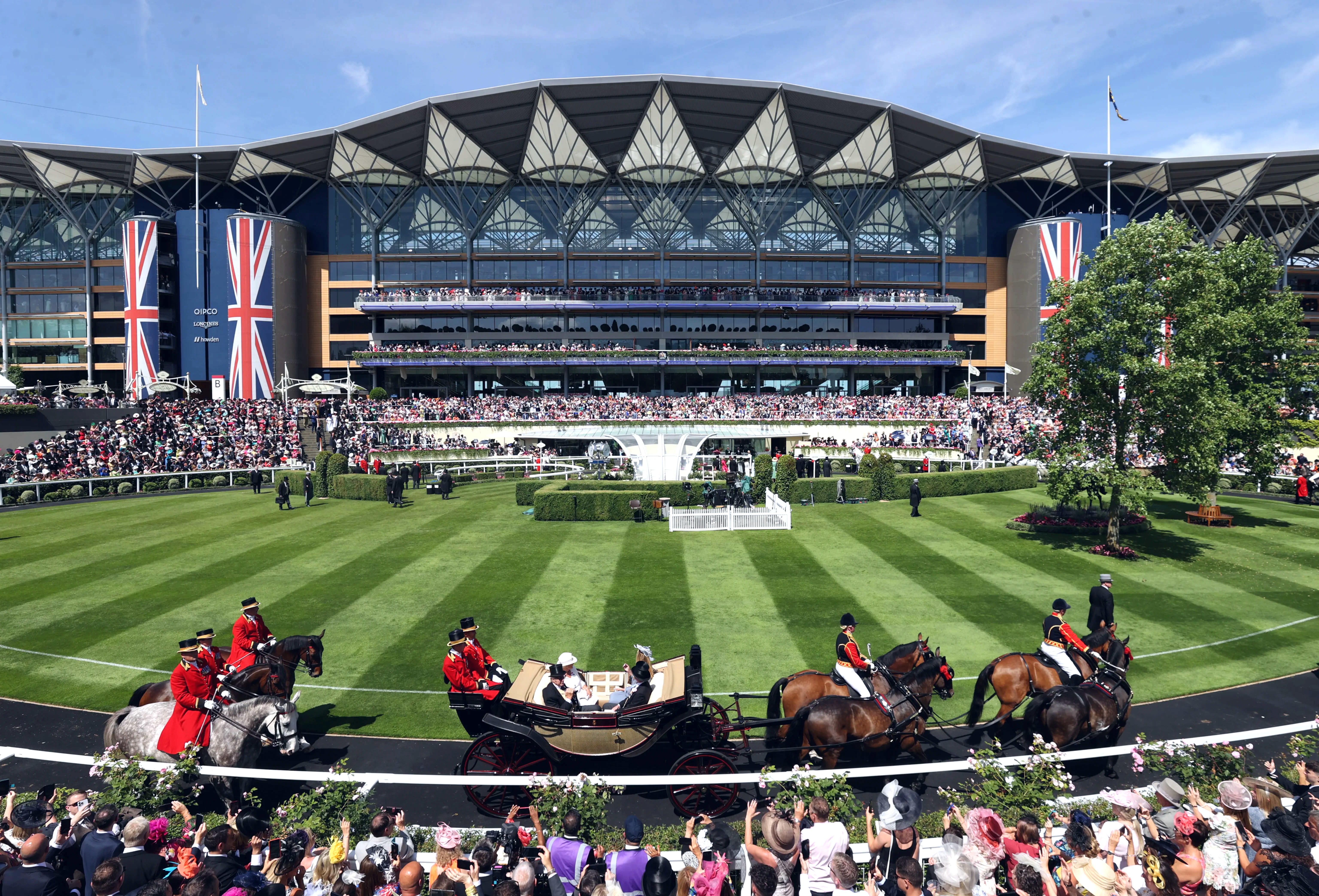 Copilot said:  Alt text:  Wide view of a racecourse with a large grandstand decorated with Union Jack banners in the background. A horse-drawn carriage and mounted riders travel along the track in the foreground, while spectators fill the stands and line the course. Manicured green lawns, a central fountain, and trees frame the scene under a partly cloudy sky.