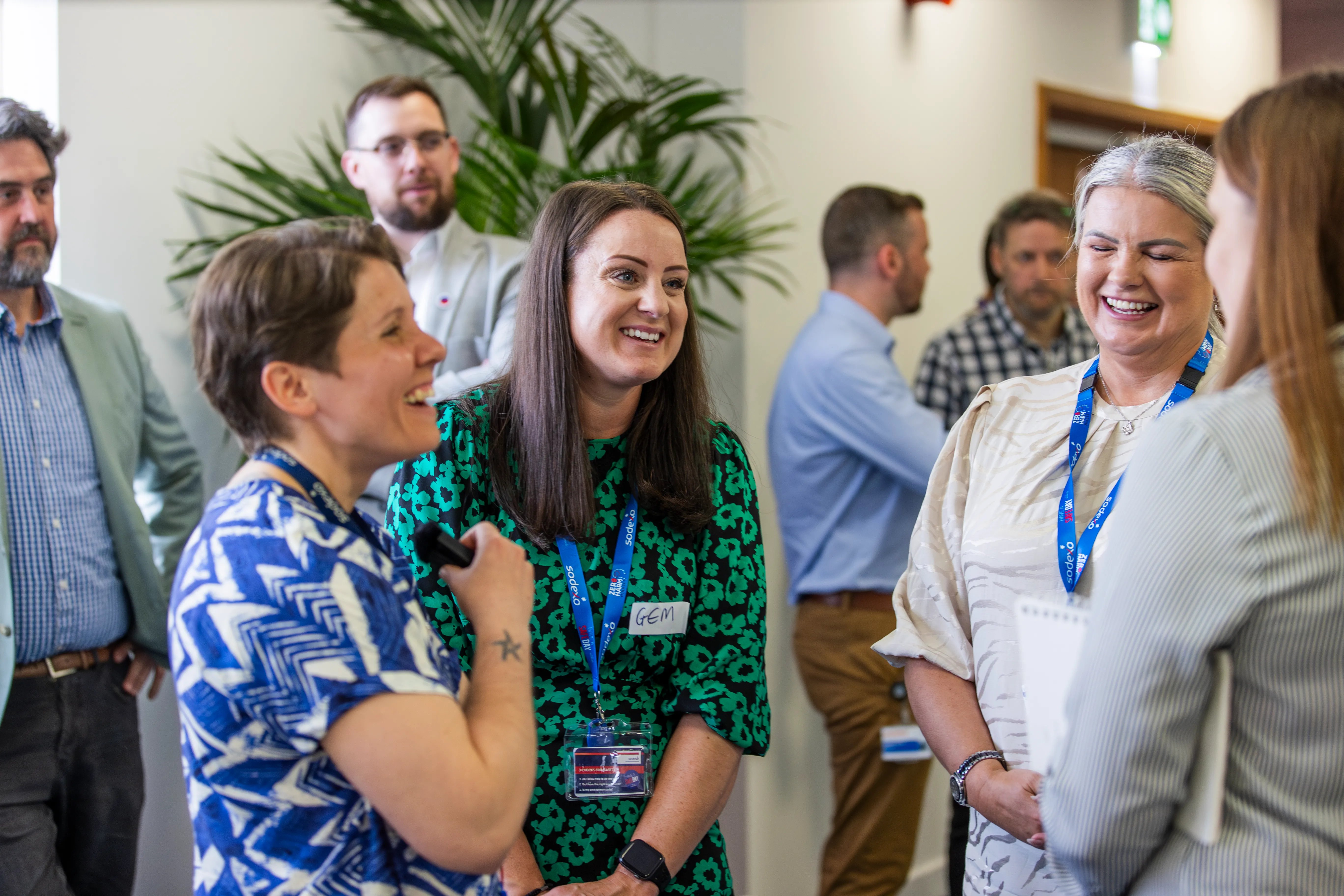 Colleagues standing together in a bright office space, talking and networking during a workplace event, with plants and other team members visible in the background.
