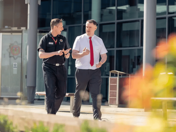 Two uniformed professionals walking and talking outside a modern building with large glass windows, with landscaped greenery in the foreground.