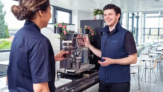 Person serving coffee at a modern café counter while showing a tablet screen, with an espresso machine and potted flowers visible in a bright indoor setting.