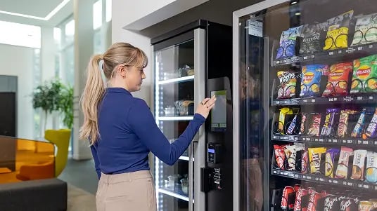 Person using a touchscreen vending machine beside a refrigerated smart fridge stocked with snacks and drinks in a modern workplace setting.