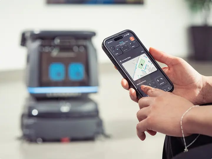 Close-up of a mobile phone controlling an autonomous cleaning robot via an app, demonstrating smart facilities management technology.