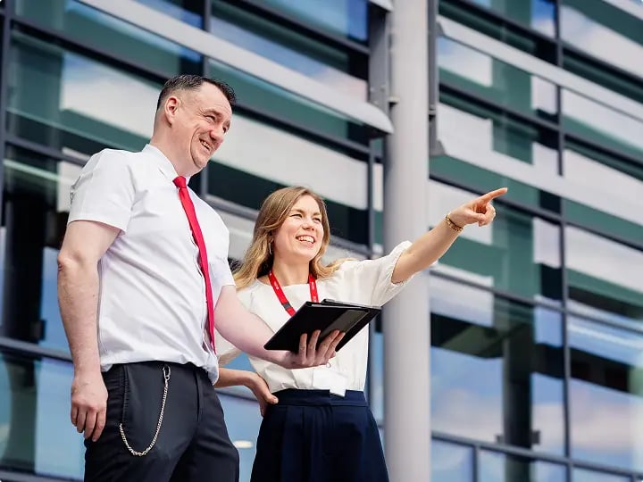 Two professionals outside a modern public building reviewing information on a tablet, with one pointing ahead, representing collaborative decision-making.