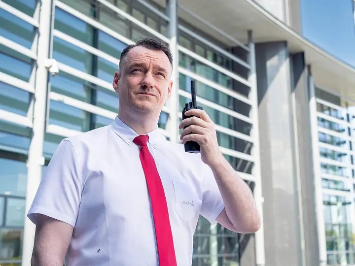 Facilities staff member using a two-way radio outside a government building, highlighting on-site security and operational coordination.