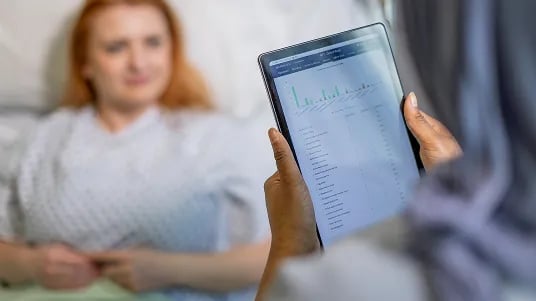Healthcare employee using a tablet to review service performance data while speaking with a patient in a hospital ward.