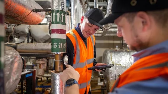 Facilities management employees inspecting building services equipment and reviewing maintenance information on a tablet in a plant room.