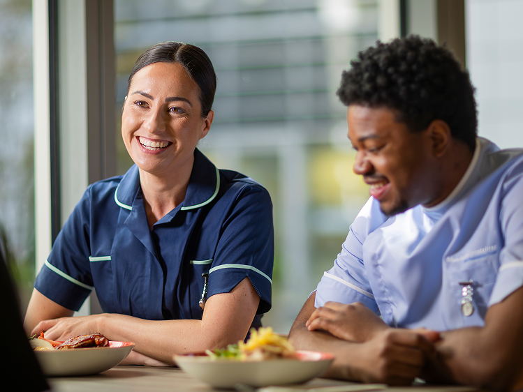 Healthcare colleagues sharing a meal and conversation in a hospital dining space, supporting workplace wellbeing and team experience.