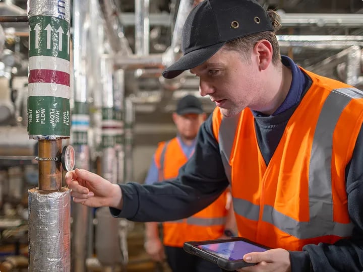 Facilities management operative inspecting building services infrastructure and recording maintenance information on a tablet.