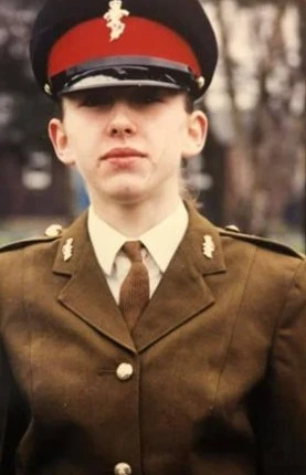 Person in formal military uniform with a peaked cap featuring a red band, standing outdoors with a blurred background.