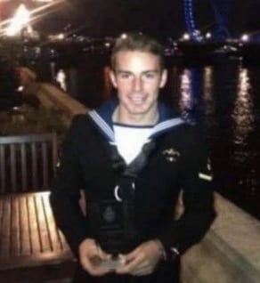 Naval service member in uniform standing by a riverside walkway at night, with city lights and reflections on the water in the background.