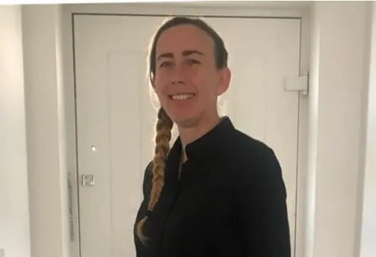 Person standing indoors by a doorway, smiling, wearing a dark top.