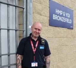 Person in work uniform standing outside a building next to a sign reading “HMP & YOI Bronzefield.”