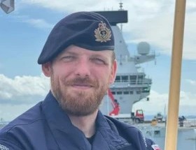 Naval service member in uniform standing on a ship with the sea and vessel equipment in the background.