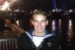 Naval service member in uniform standing by a riverside walkway at night, with city lights and reflections on the water in the background.