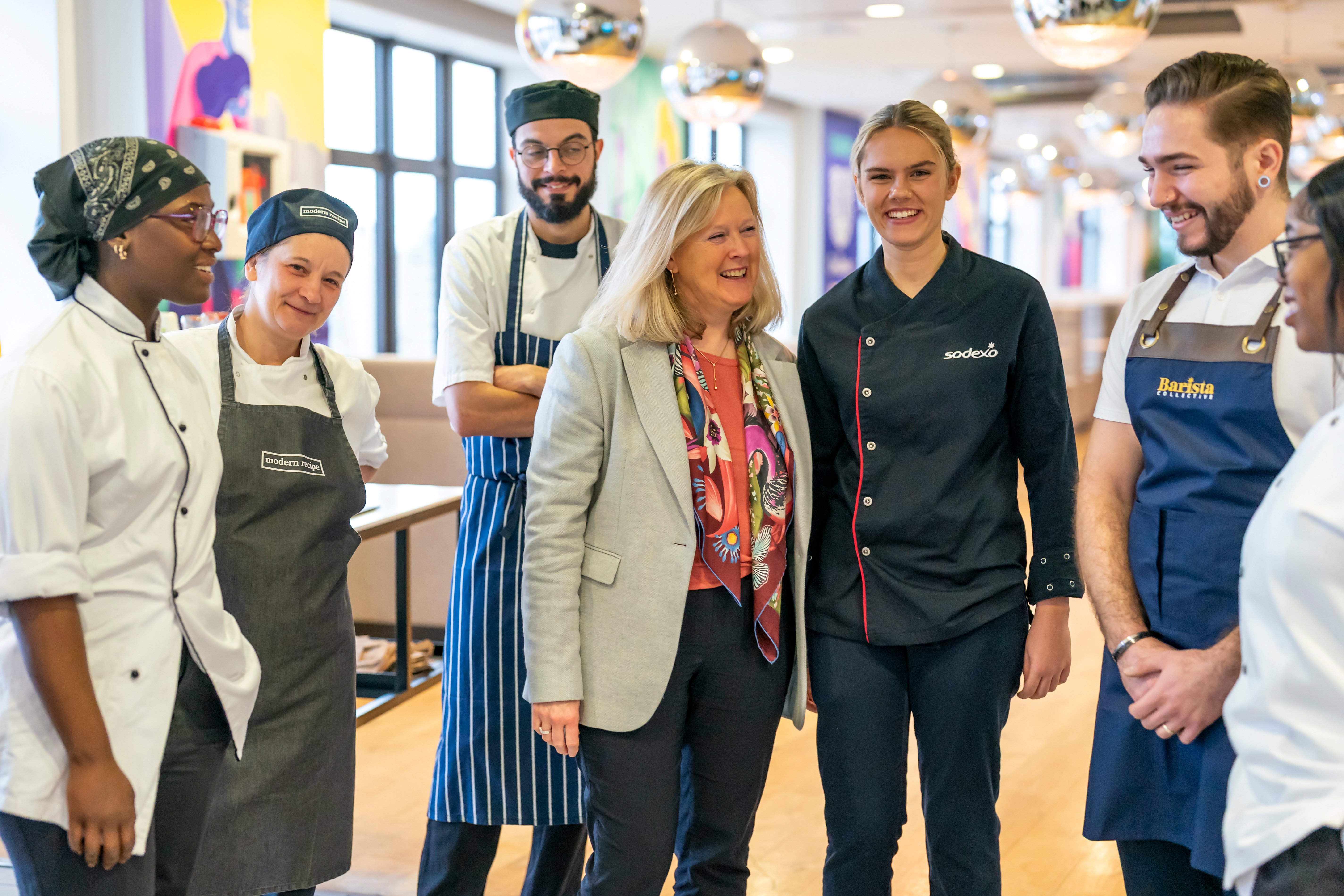 A small group of adults wearing chef and hospitality uniforms stand together in a bright indoor space, arranged in a loose semi-circle, with worktables, windows, and hanging spherical lights visible in the background.