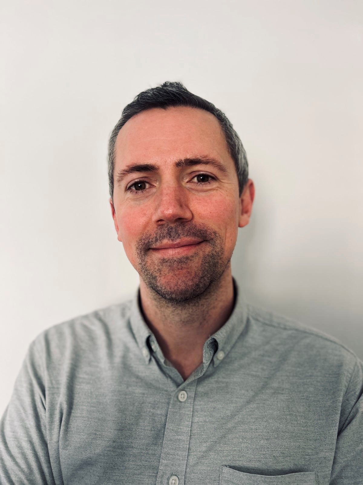Head-and-shoulders portrait of a person facing the camera, wearing a light grey button-up shirt, positioned against a plain off-white background.