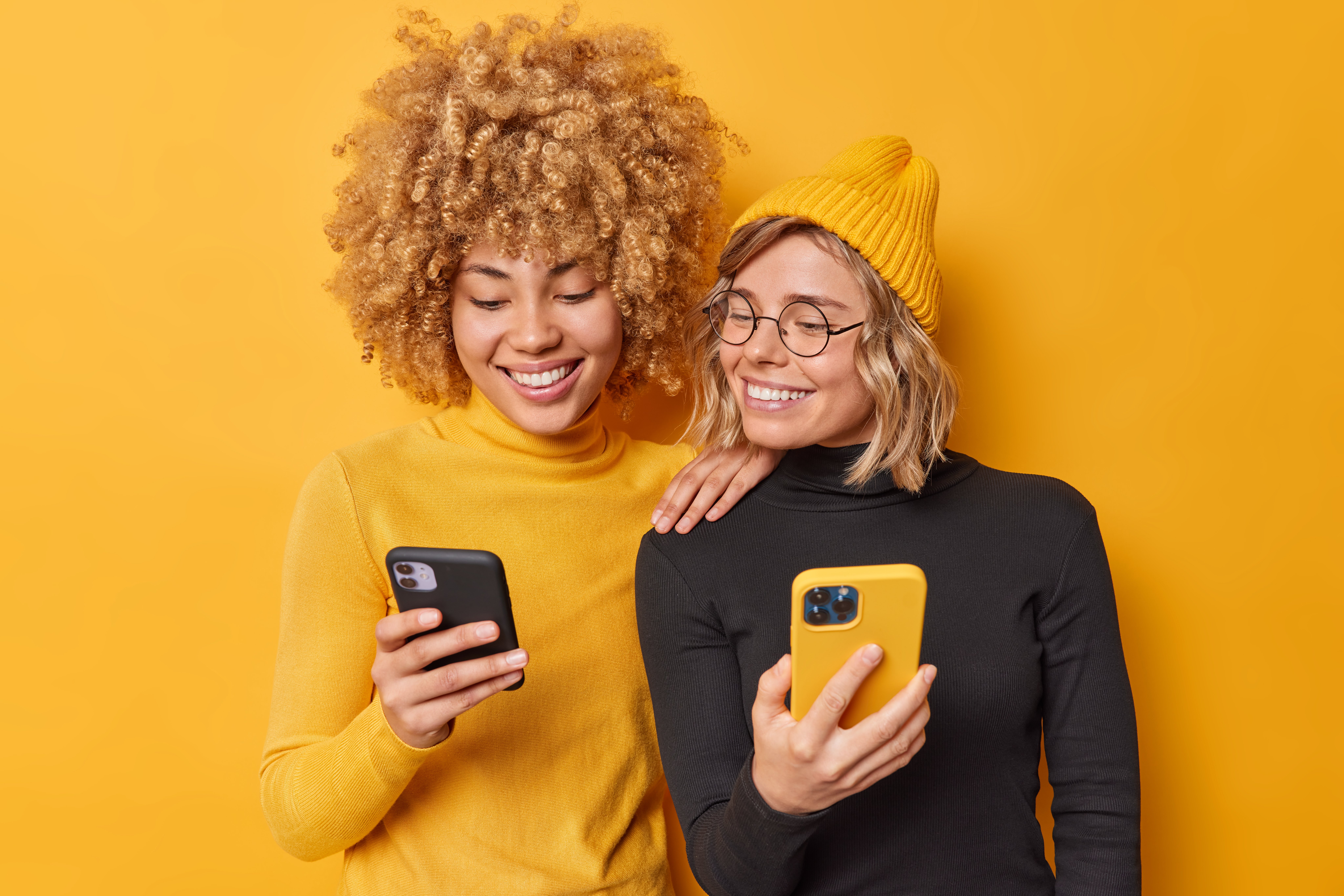 beautiful-two-female-friends-use-mobile-phones-browse-internet-type-sms-message-dressed-casual-turtlenecks-smile-gladfully-isolated-vivid-yellow-background-watch-video-via-media-app