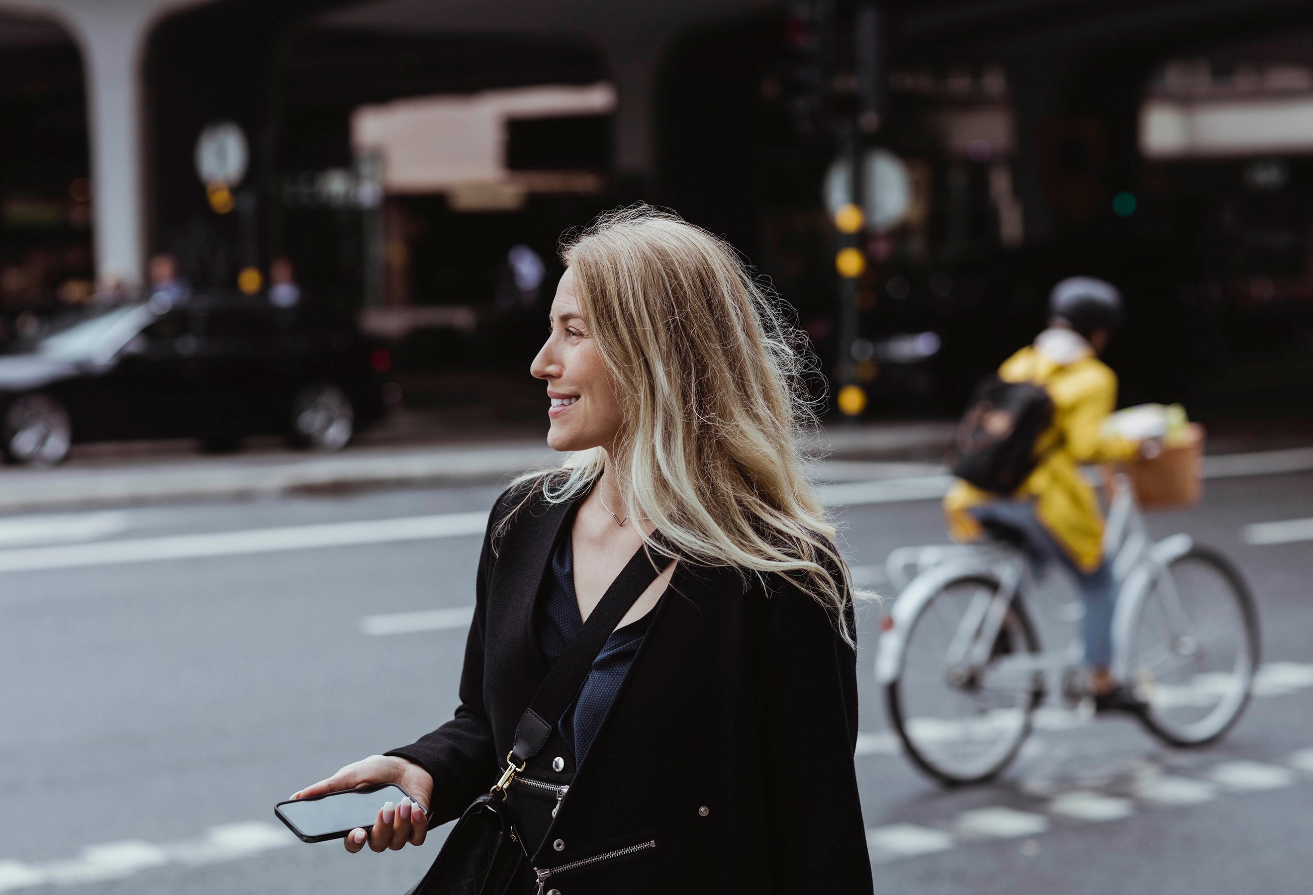 Dame med mobil i handen ute på gaten. Syklist og biler i bakgrunnen.