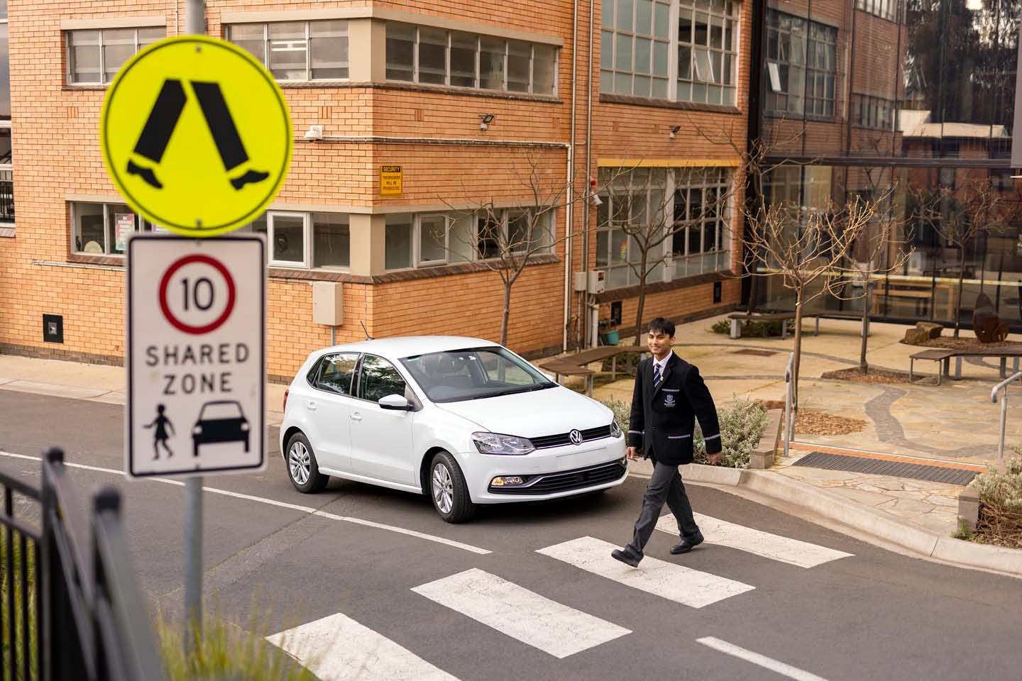 Photograph of a school student walking across a pedestrian crossing in front of his school, with a white hatchback car stopped to let him pass