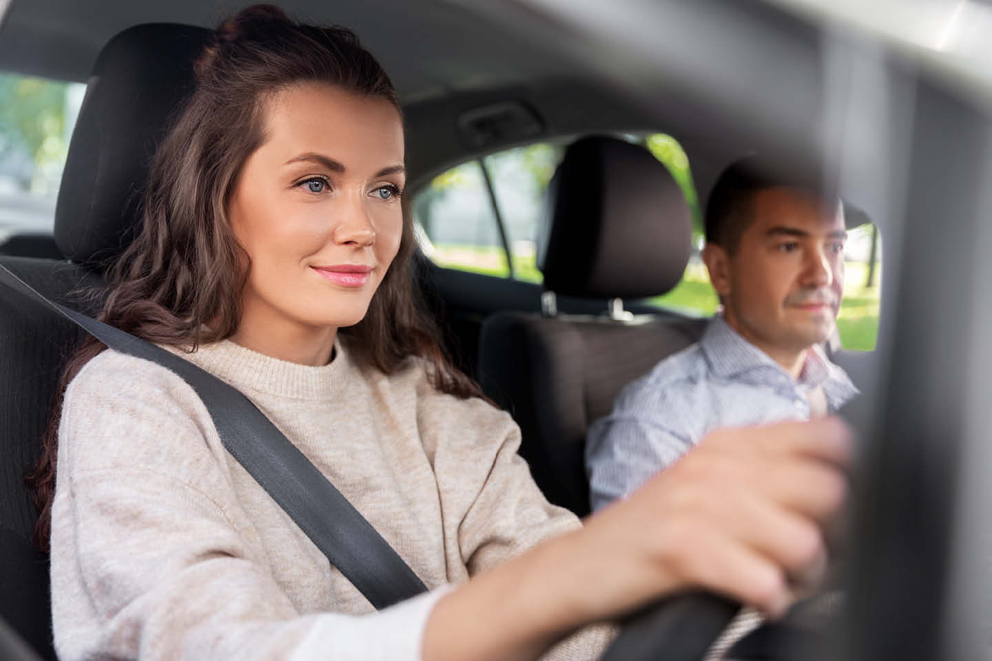 Photograph of a young woman driving a car and wearing a seatbelt, with a man in the passenger seat