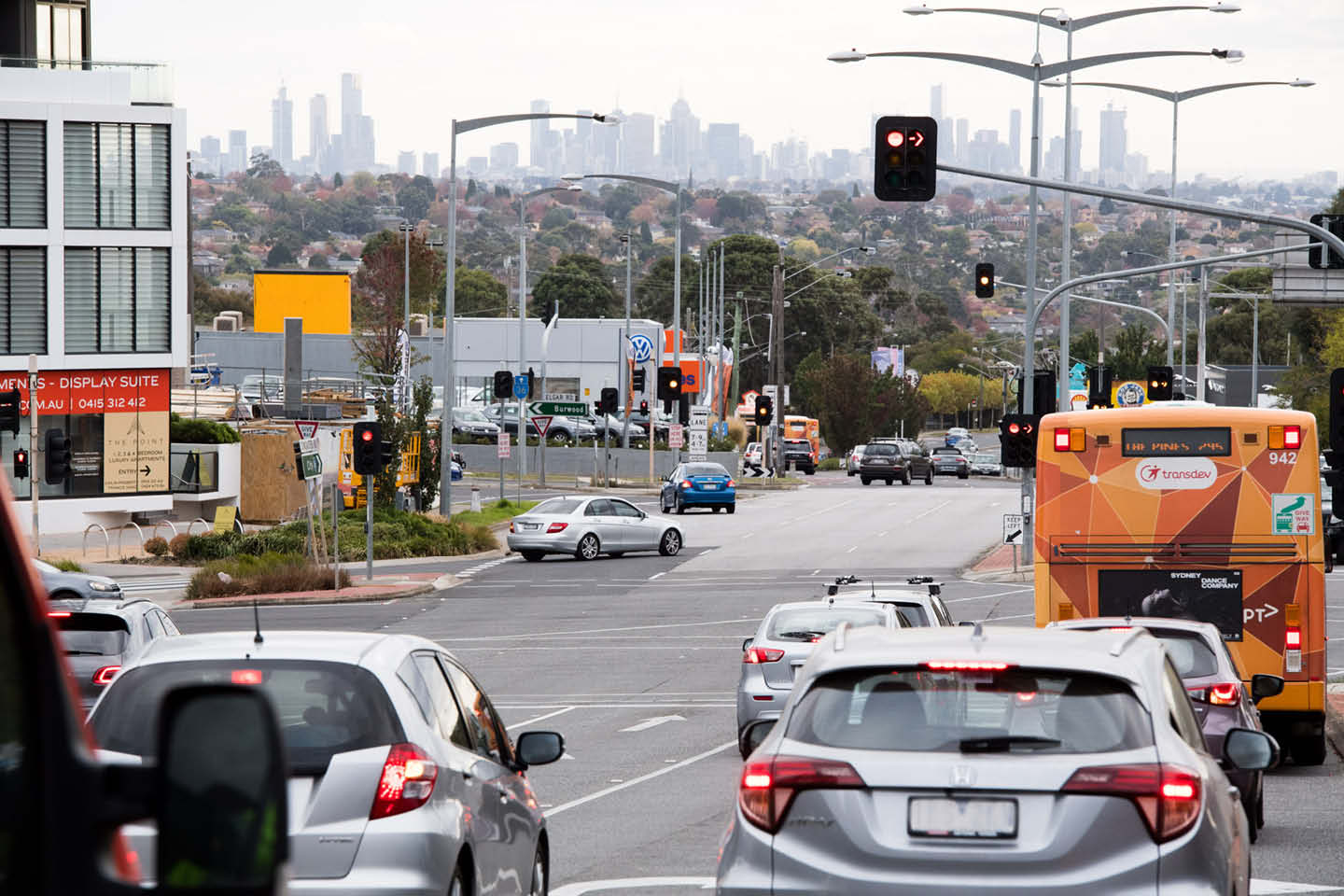 Photograph of cars pulling up to a red light at an intersection, Melbourne city skyline in the distance