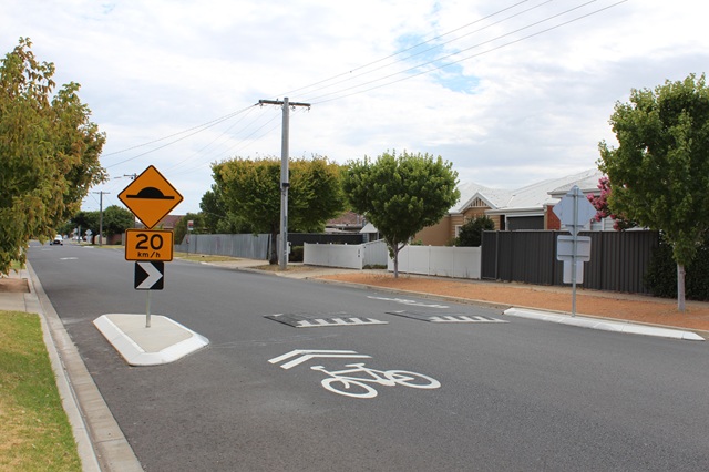 Image of cycling marking on a road next to a speed hump. 20 km/h yellow and black sign also shown on the speed hump.