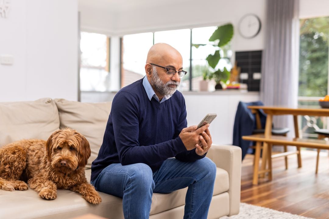 Man sitting on a couch looking at his phone. There's a dog on the couch next to him.