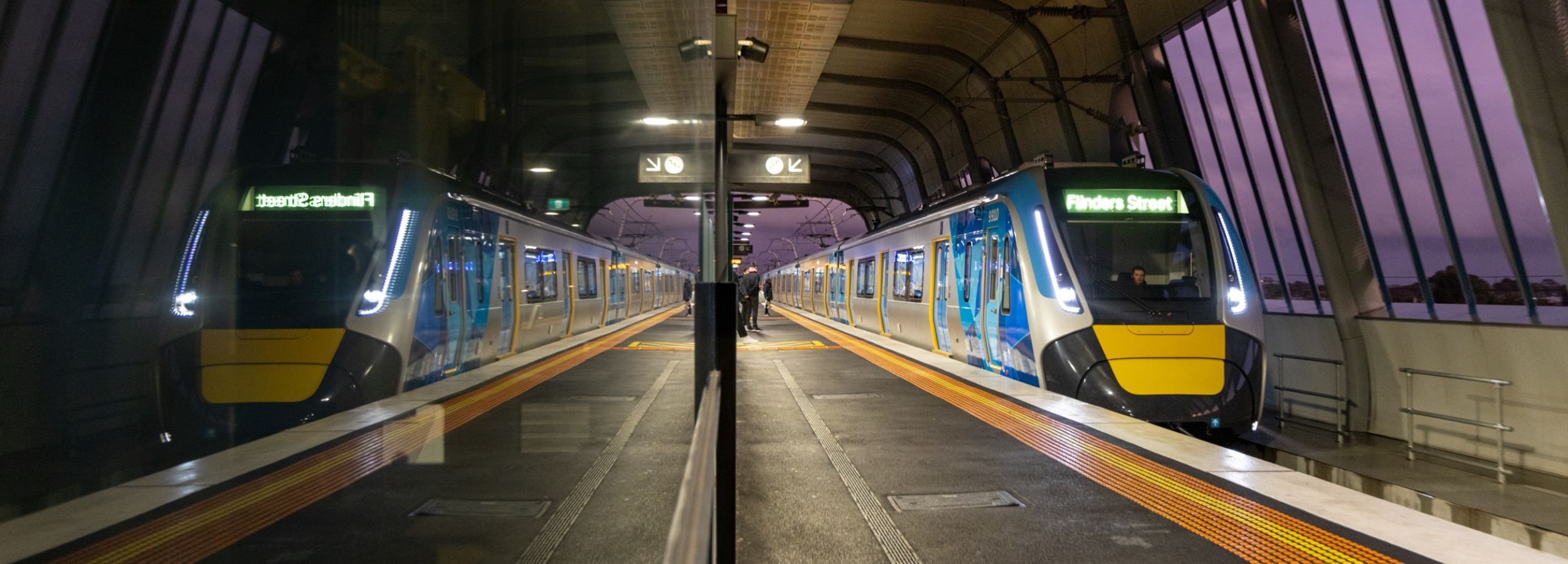 Metro train at station platform. It is dusk and the sky is purple. The image is reflected in a glass panel.