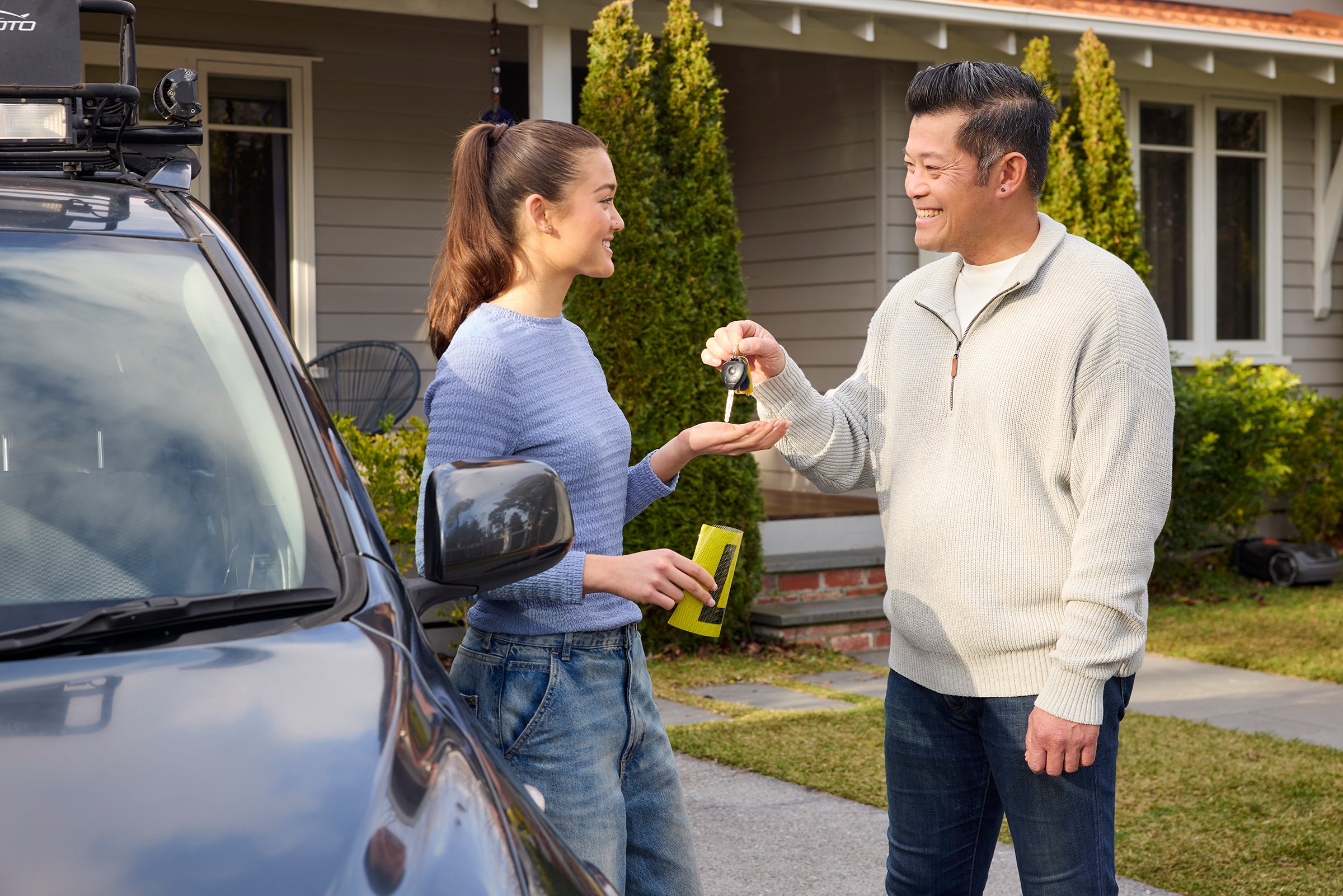Woman holding a yellow L plate leaning on a car being handed a set of car keys by a man