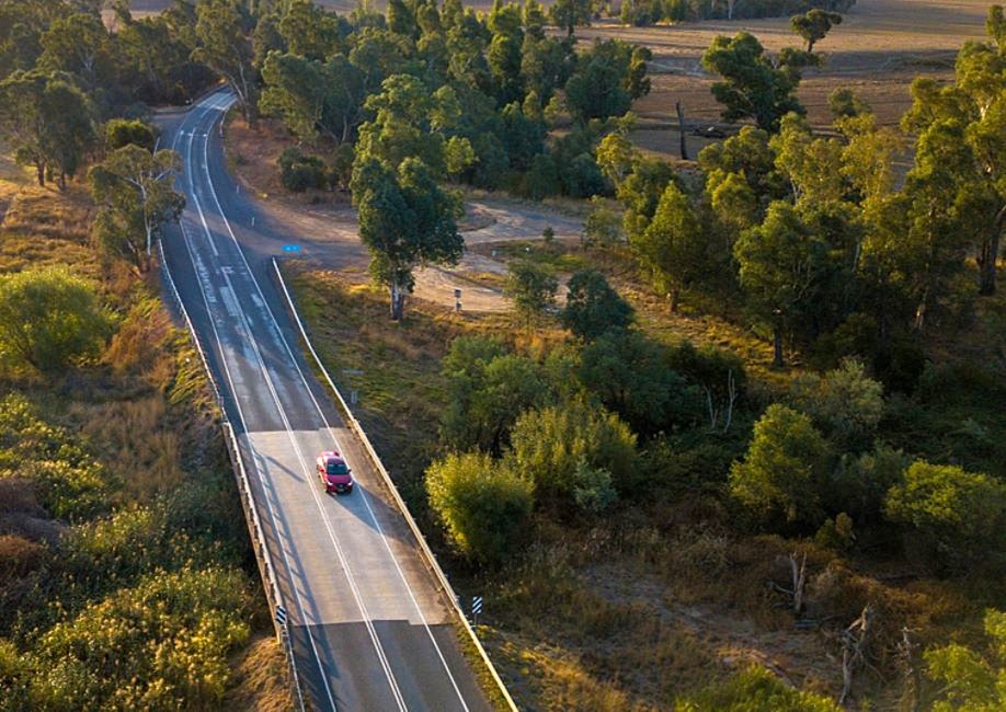 Photo of a red car driving along a country road surround by trees.