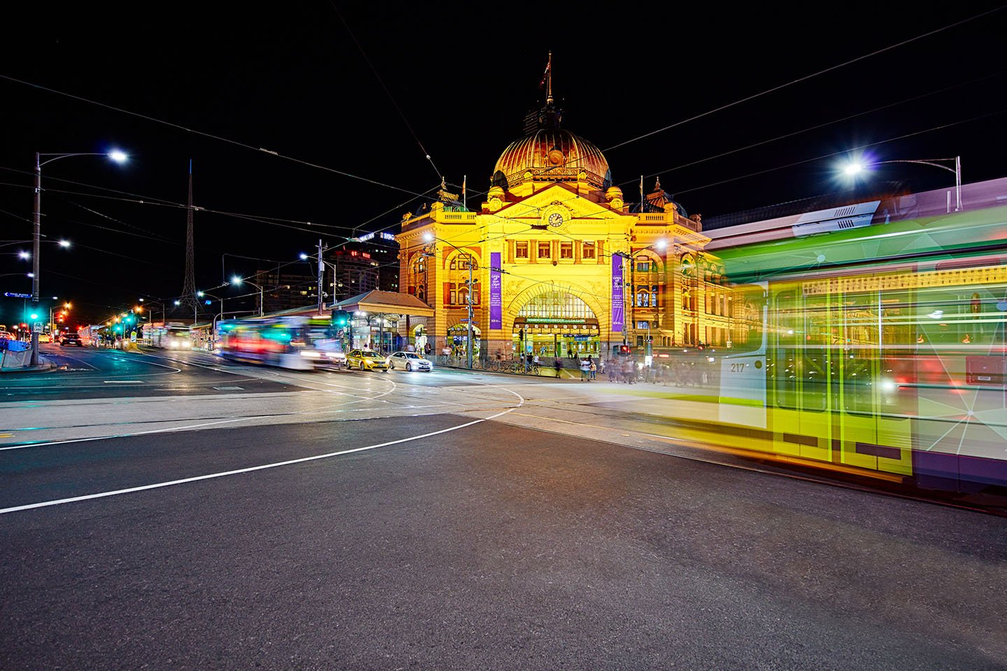 Flinders Stret station at night with trams passing by on both sides of the road.