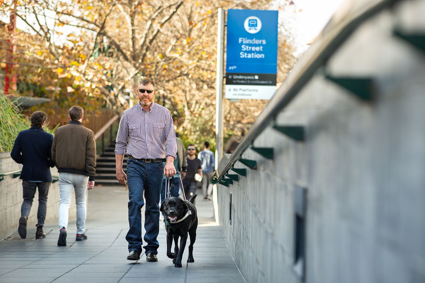 A visually impaired man with his guide dog walking along. There is a sign for Flinders Stree train station behind him.
