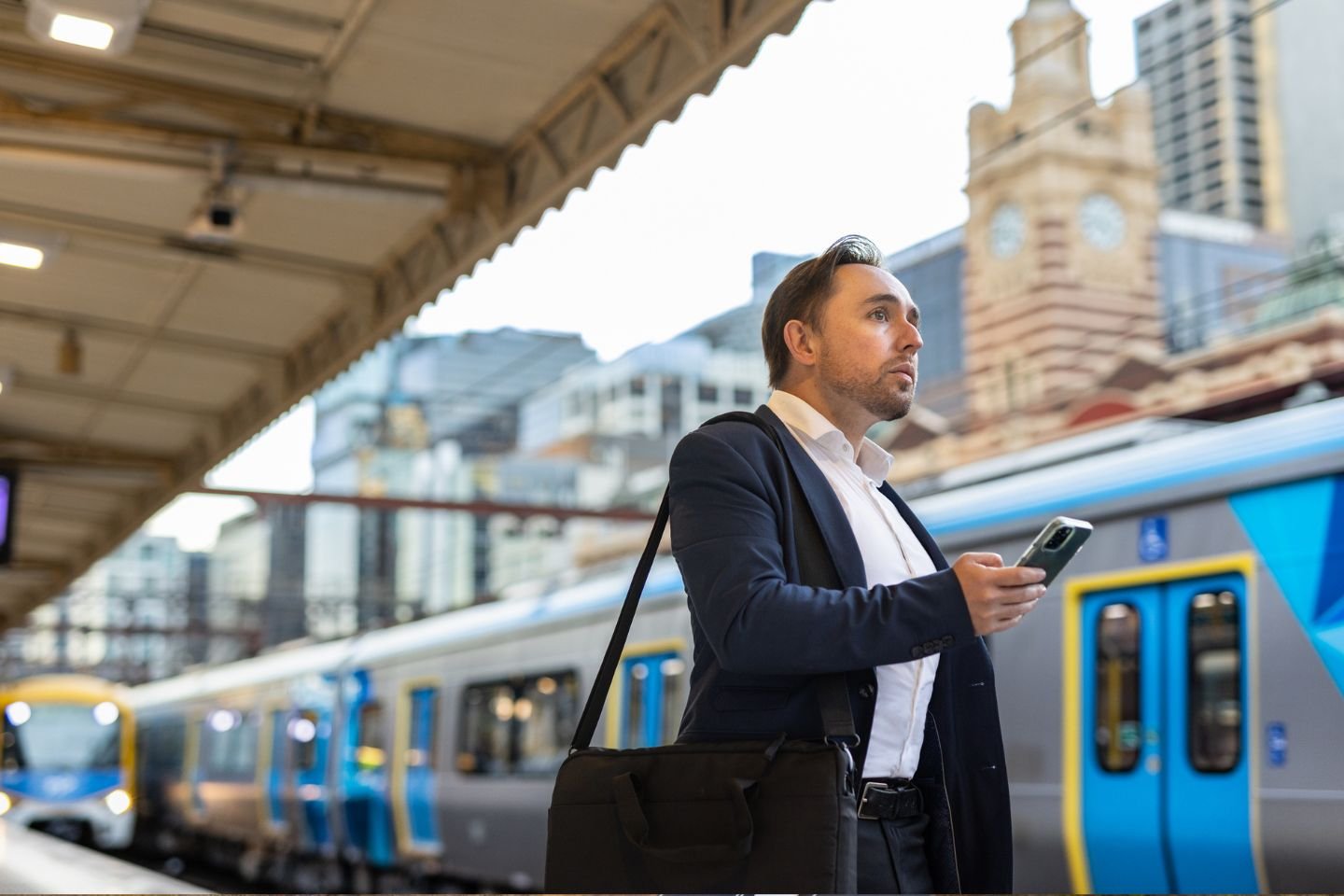 A man in a business suit standing on a platform at Flinders Street Station. He's holding his mobile phone and there's a train at another platform in the background.