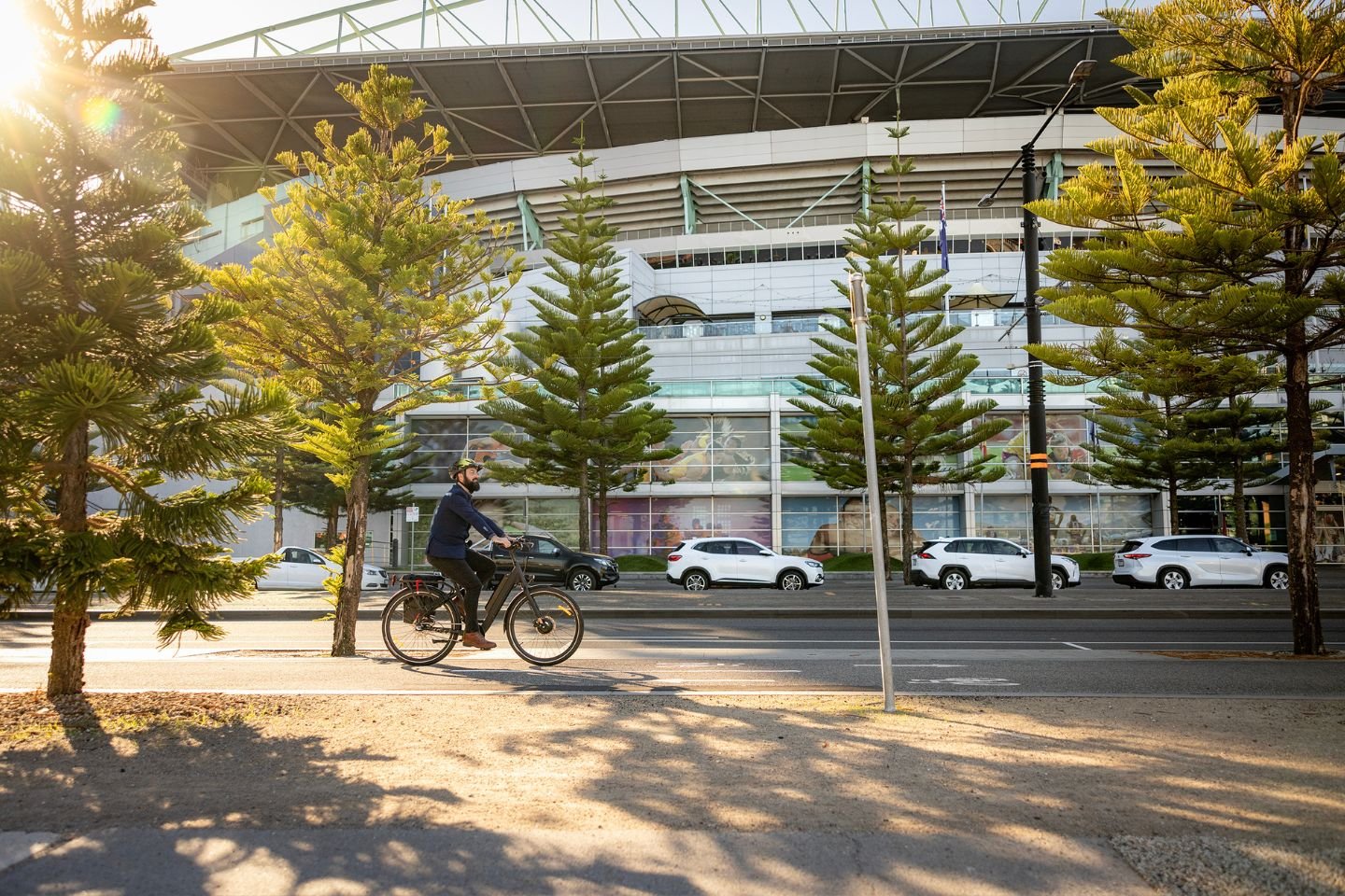 Man on an e-bike riding past a stadium. He's on an off-road shared path. There are cars parked on the other side of the road.