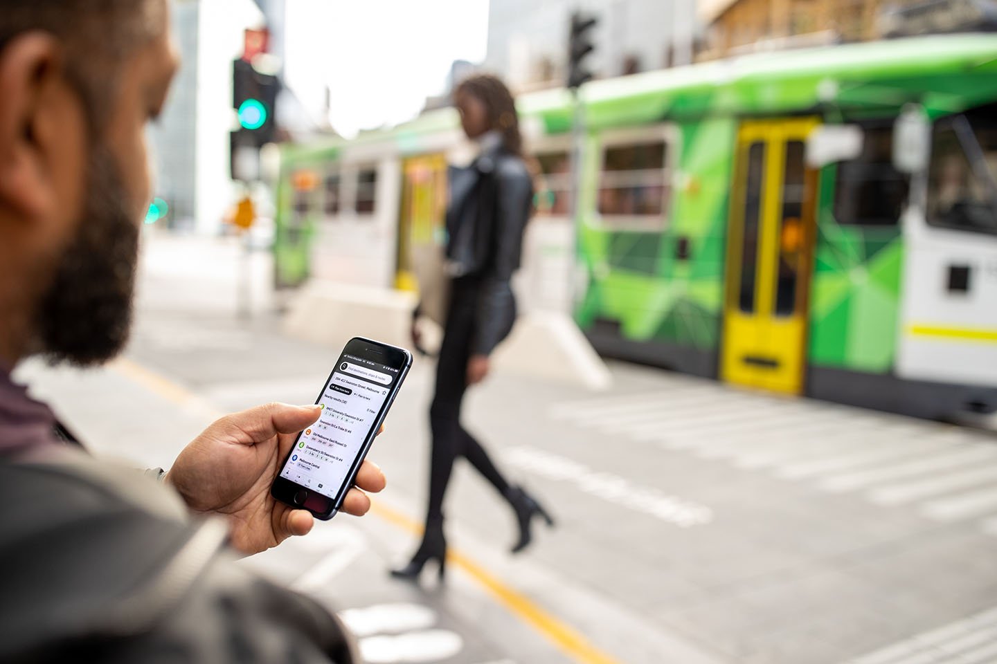 A man looking at journey planner on his mobile phone at a tram stop. A woman is walking in the background.