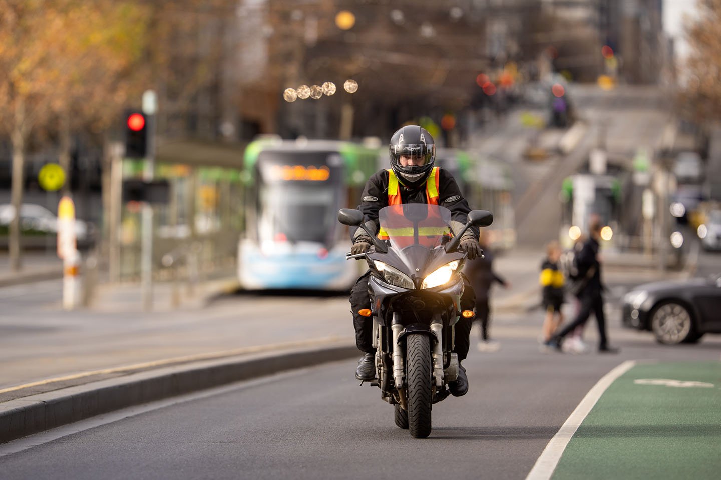 Man riding a motorbike. There is a tram and people crossing the road in the background.