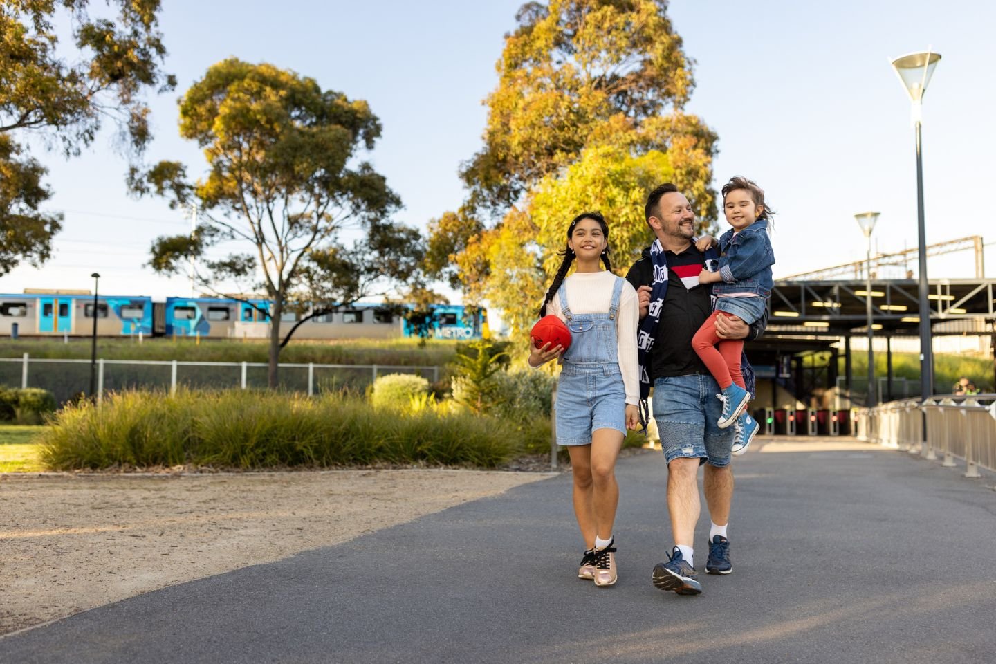 A man wearing a footy scarf walking with a teenage girl and carrying a young girl. There's a train in the background. The teenage girl is carrying a football.