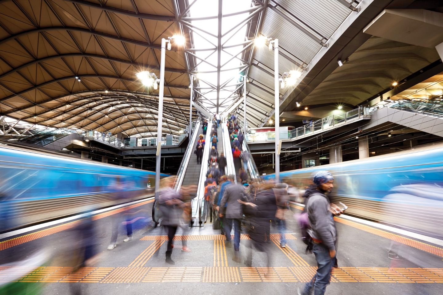 Passengers using the Souther Cross Station escalators. There are trains on each side of the platform.