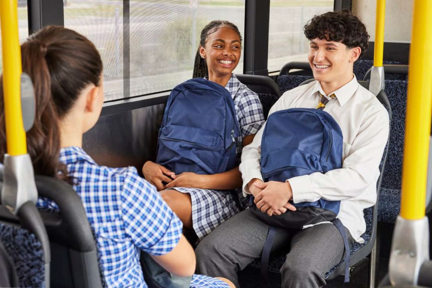 Three students in school uniforms on a bus. They're sitting down and holding their backpacks on their laps.