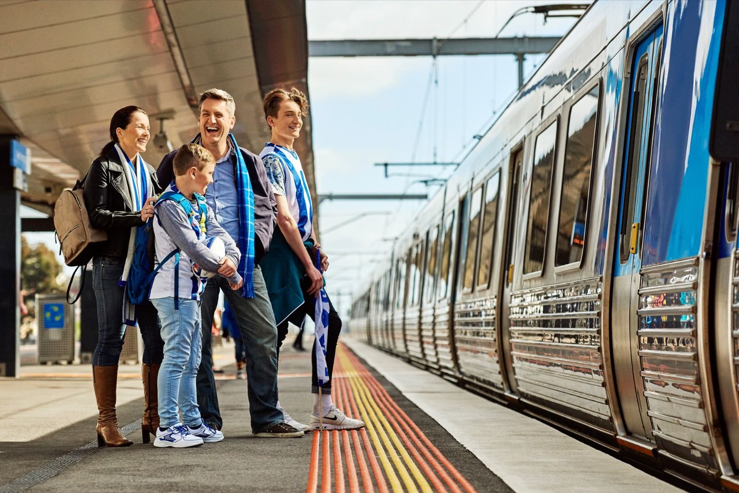 A family of a mum, dad and two boys wearing football scarfs waiting to catch a train. One of the boys has a football.