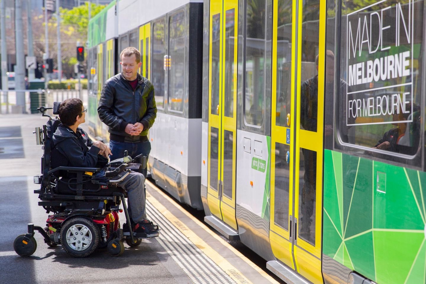 Two men waiting at a tram stop for a tram to open its doors. One man is in a wheelchair.