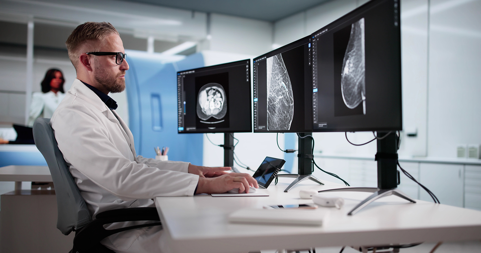 A radiologist in a white lab coat and glasses sits at a modern workstation, reviewing medical imaging on three large computer monitors.