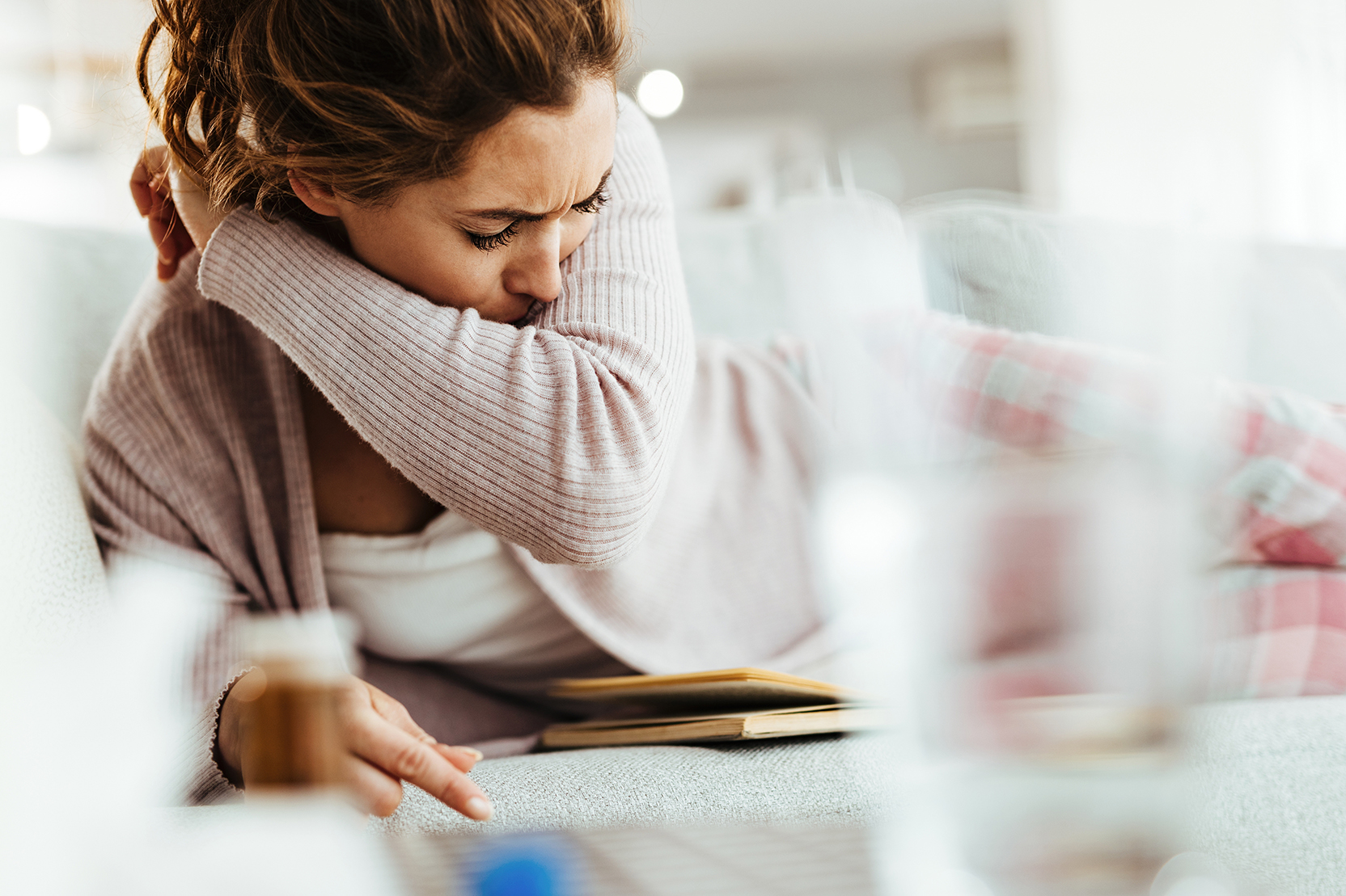 Woman coughs into her arm while reading a book on a couch. 