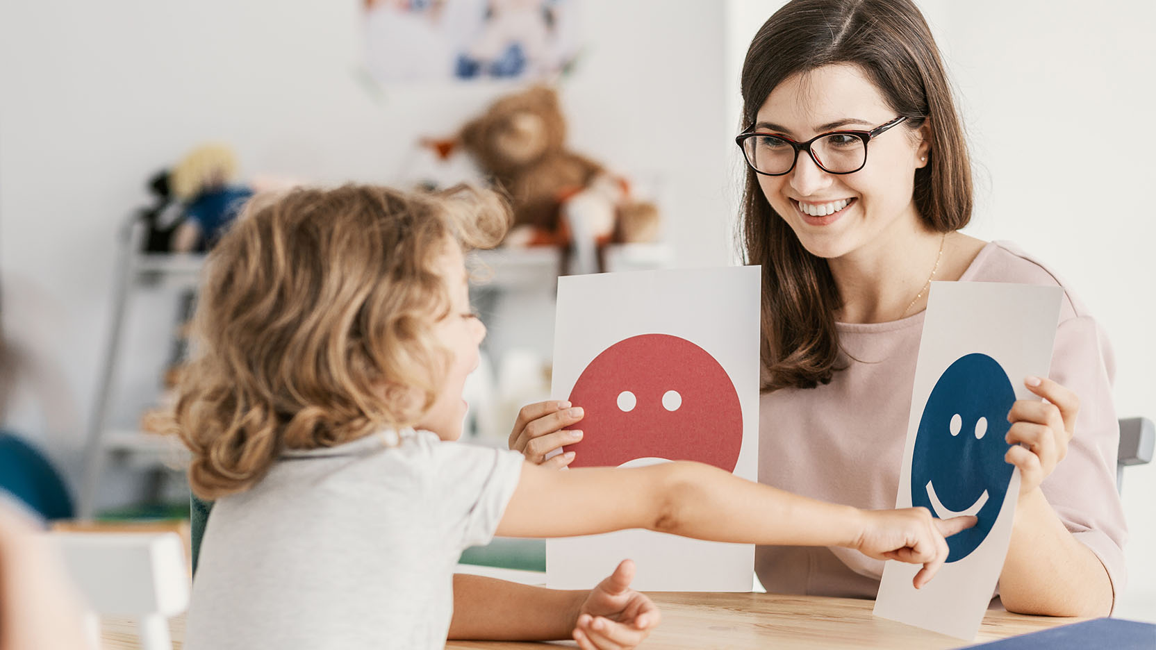A smiling psychologist shows a young child with autism emoticons during a therapy session.